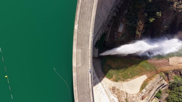 Top-down view of Baells Dam in Catalonia, Spain, showing powerful water discharge from the concrete arch dam wall into the valley below with green reservoir