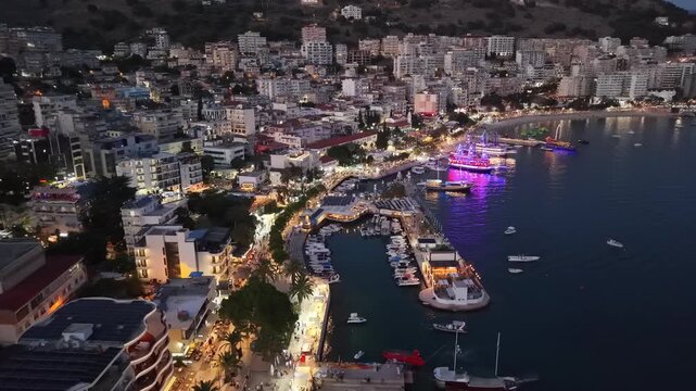 Breathtaking high angle view of Saranda waterfront nightlife at twilight, featuring glowing neon lights on cruise ships, coastal Mediterranean architecture, crowded boulevard walkway, and scenic citys