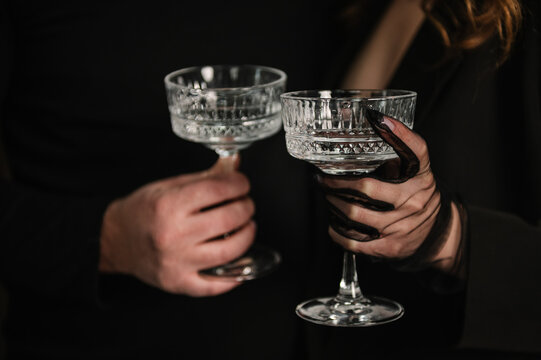 Cheers. Close-up of a man and a woman in black clothes holding elegant crystal coupe glasses. They are making a toast. Dark moody atmosphere, sophisticated celebration, evening party concept.