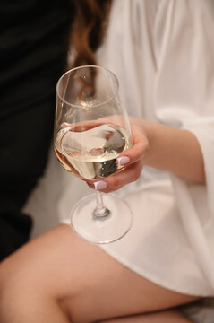 A close-up of a woman's hand holding a glass of white wine. She wears a white silk robe and has a french manicure. The setting is indoors, during a wedding morning.