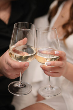 Cheers. A romantic close-up of a man's and a woman's hands clinking glasses of white wine. The setting is indoors, during a wedding morning or celebration. Soft focus background.