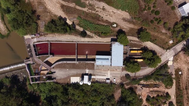 Top view of a water intake channel at a drinking water treatment plant showing deep red water dosed with potassium permanganate for pre-oxidation, flowing from adjacent river toward treatment facility