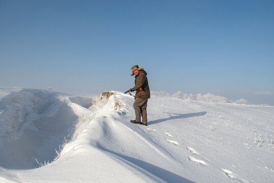 A hunter in warm clothes and a fur hat stands on the edge of a snow-covered ravine with a shotgun.
