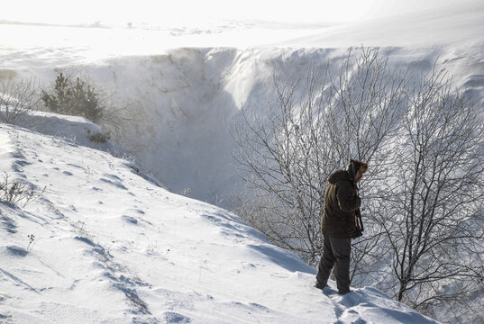 A hunter with a shotgun stands on the edge of a snow-covered ravine on a winter day.