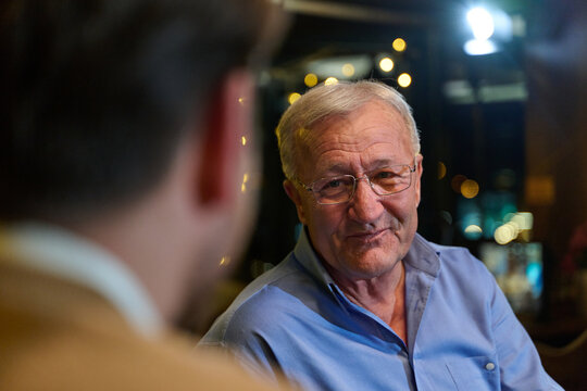 Warm Candid Portrait Of An Older Man Smiling During A Nighttime Conversation At A Restaurant