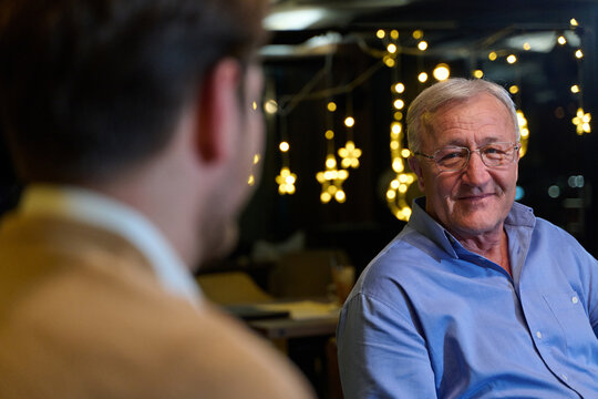 Smiling Senior Man Conversing With Younger Relative In Cafe With Festive Christmas Lights