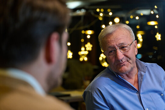 Elderly Man Conversing With Younger Person In Cozy Cafe With Festive Bokeh Lights