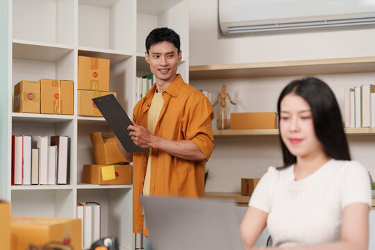 Asian small business owner holding clipboard looking at female colleague using laptop