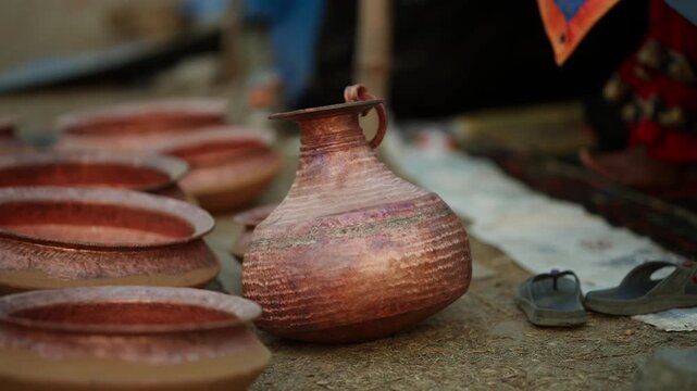 Rows of hand hammered copper bowls and a traditional water jug with a handle sit on the ground beside a pair of sandals outdoors