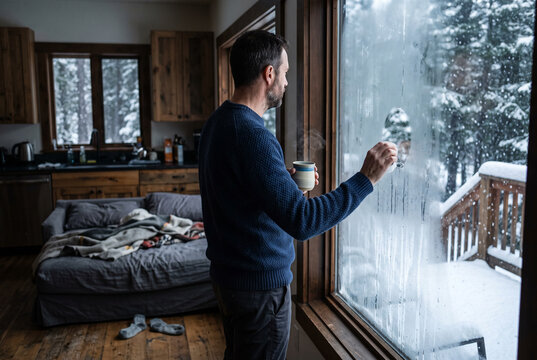 Man in a rustic cabin looking out a frosted window at falling snow holding a coffee mug for cozy winter morning lifestyle background and home retreat design