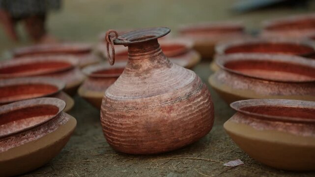 A traditional copper water jug with a small handle stands centered among rows of hand hammered copper bowls arranged neatly on the ground outdoors