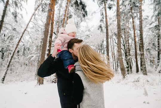 Parents kissing during winter walk, father carrying little daughter on shoulders in snowy forest. Happy family love and emotions. Closeup.