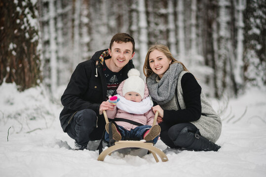 Happy parents sitting beside little daughter on sled in snowy forest. Family smiling during winter walk, enjoying time together. Cozy family leisure in nature. Close up.