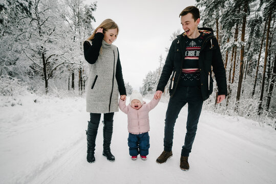 Happy parents laughing, leading child by hands along snowy road in winter forest. Toddler emotionally reacting to walk. Family idyll, winter adventures and active leisure outdoors.