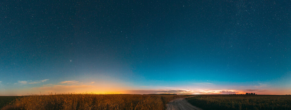 Night Starry Sky With Glowing Stars Above Countryside Landscape. Light Cloudiness Overcast Above Rural Field Meadow And Country Road In Summer. Panorama, Panoramic View