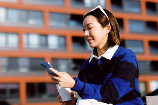 Young asian woman holding a coffee cup and interacting with her smartphone while sitting outside a modern building