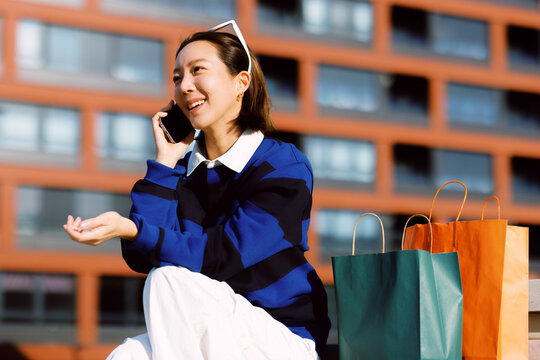 Young asian woman smiling and communicating on her mobile phone outdoors sitting next to colorful shopping bags