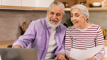 Fototapeta premium Smiling Elderly Couple, Man And Woman, Engaged And Enjoying Time Together Using Laptop In Cozy Home Setting, Sharing Joyful Moments. Sweet Affection And Companionship Concept.