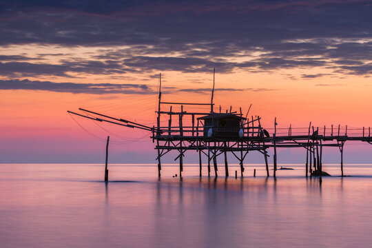 View of the Costa dei Trabocchi, a traditional wooden fishing structure on stilts over the calm sea during a vibrant sunset in Marina di San Vito, Abruzzo, Italy.