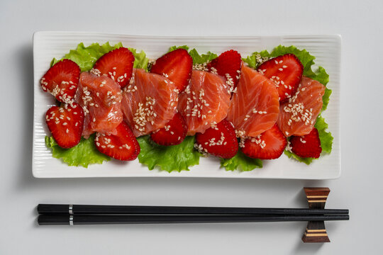 Salmon sashimi appetizer with strawberries and sesame. Gourmet salmon carpaccio with red strawberry slices on green lettuce leaves with sesame seeds served with chopsticks on white background