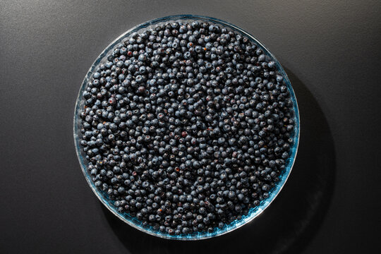 Fresh wild blueberries in round glass bowl, closeup. Top view of circular transparent plate filled with large amount of ripe dark blue forest blueberries placed on black background with shadows