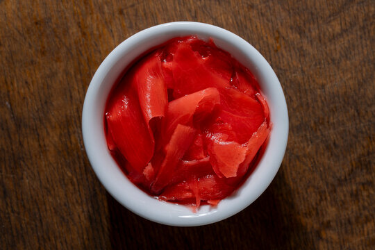 Pickled pink ginger slices in white ceramic bowl, closeup. Top view of small white round bowl filled with bright pink pickled ginger gari placed on rustic dark wooden table background