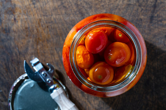 Open jar of pickled tomato on wooden background, closeup. Top view of an opened glass jar with pickled red and yellow cherry tomatoes in brine next to metal lid and can opener on rustic wooden table
