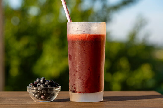 Fresh blueberry smoothie in tall glass outdoors. Chilled dark berry smoothie or juice in frosted glass with straw next to fresh blueberries on wooden table with blurry green garden background