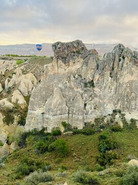 View of Cappadocia with its iconic fairy chimney rock formations, green valley slopes, and hot air balloons floating in the distance under a cloudy sky in Turkiye.