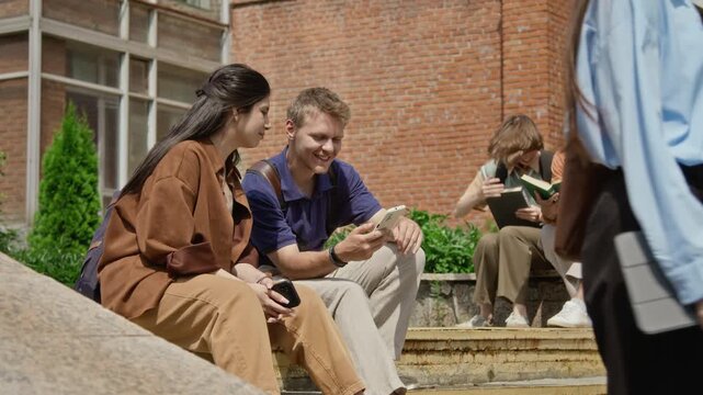 Caucasian young man and Hispanic girl sitting on campus steps, looking at smartphone. Ideal for promoting higher education apps and university student life campaigns
