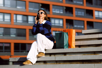 Fototapeta premium Young woman wearing sunglasses sitting on urban stairs talking on her smartphone with colorful shopping bags beside her