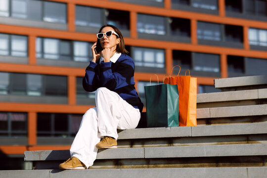 Young woman wearing sunglasses sitting on urban stairs talking on her smartphone with colorful shopping bags beside her
