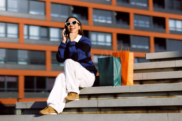 Fototapeta premium Smiling woman sitting on city steps resting with shopping bags while having a cheerful mobile phone conversation