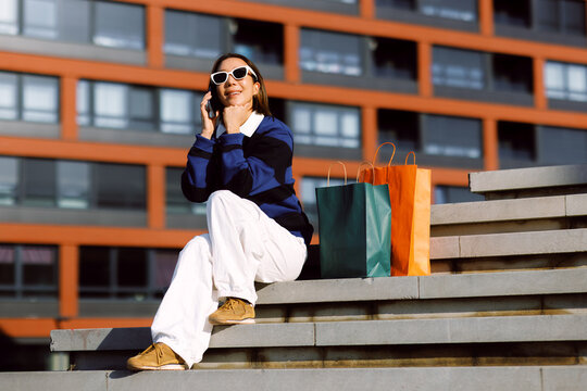 Smiling woman sitting on city steps resting with shopping bags while having a cheerful mobile phone conversation