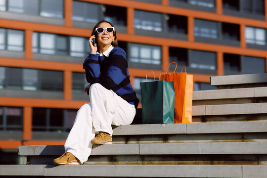 Smiling woman wearing sunglasses sitting on urban stairs talking on smartphone with colorful shopping bags beside her