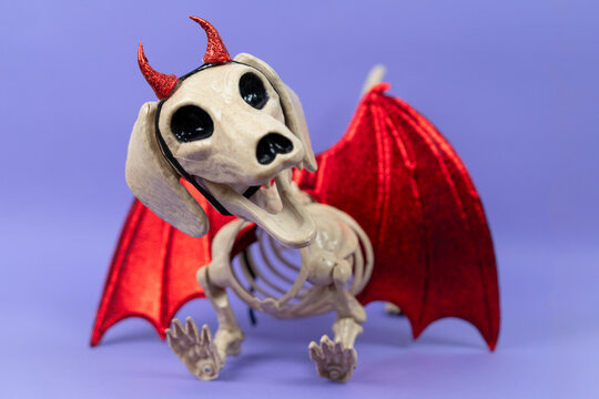 A spooky dachshund skeleton prop wearing red glitter devil horns and crimson bat wings against a purple backdrop, a scary Halloween display concept for eerie seasonal marketing.