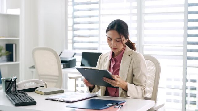 A serious Asian businesswoman is intently searching through a stack of documents while sitting at her desk in her office.