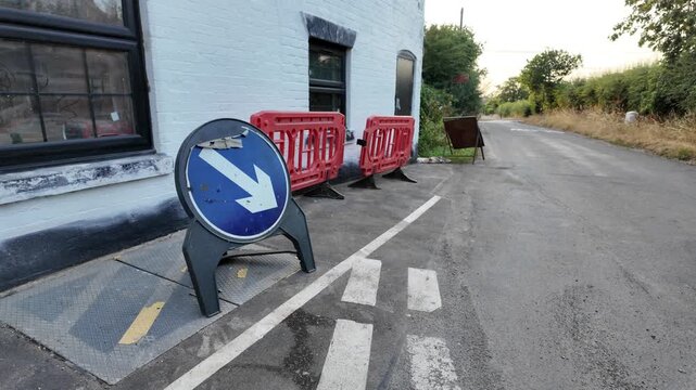 Roadside sign directs traffic toward a closed route. Red barriers block access near a building. Pedestrian crossing marks the restricted area. No people are visible in the scene