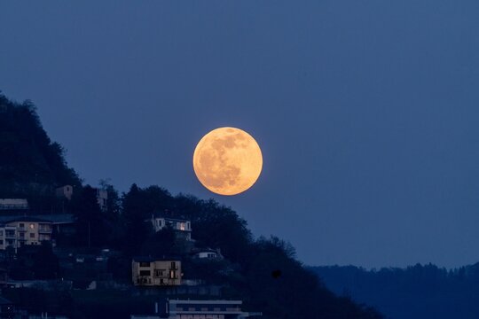 View of a large full moon rising over a hillside with residential buildings at twilight in Lugano, Switzerland.