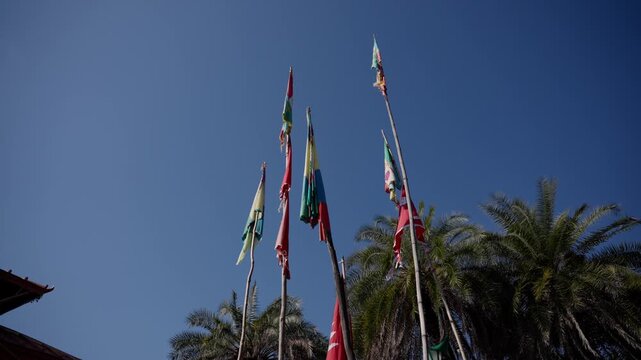 Colorful religious prayer flags fluttering atop tall bamboo poles against a clear blue sky with palm trees and a temple roof edge