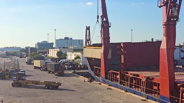 An elevated daytime shot of a large geared cargo ship docked at a port, using its onboard red cranes to lift a shipping container from a busy terminal yard filled with trucks.