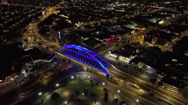 Wide nighttime drone orbit reveals Avenida Aguascalientes Sur across much of its route, with flowing traffic, the blue-lit Bicentenario Bridge and the extensive cityscape of Aguascalientes, Mexico.