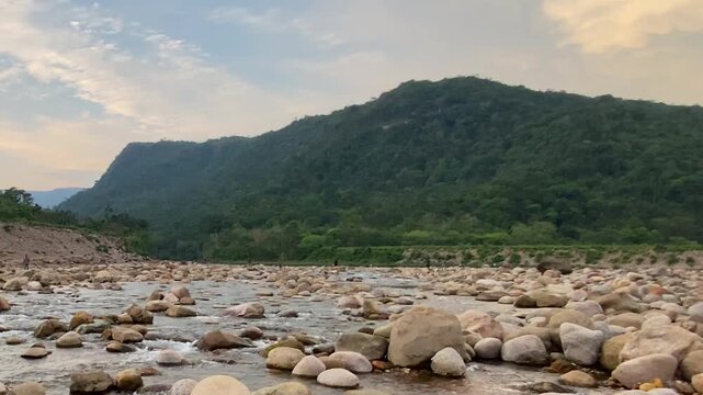 Mountain river stream flowing over rocks with a large reddish boulder in foreground, framed by Meghalaya hills under a cloudy sky at Bichanakandi in Sylhet, Bangladesh. Wide low-angle view