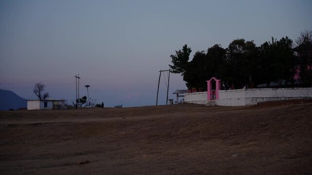 Quiet dusk scene of a rural village with a pink temple gate white boundary wall and tall trees beneath a fading blue evening sky, Farwest Nepal