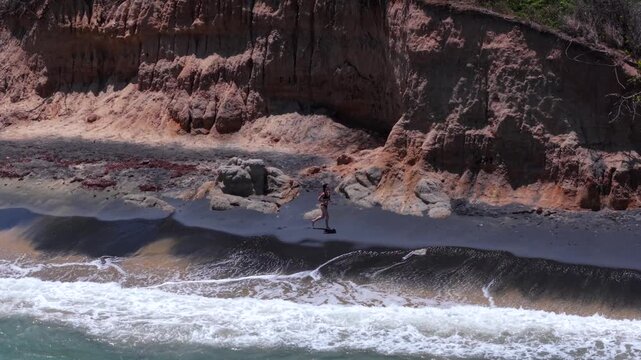 A woman runs along the black sand of Playa Negra, framed by rocky cliffs and turquoise waves in Vieques, Puerto Rico, telephoto tracking shot