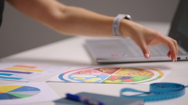 Side view of a female nutritionist working on a laptop while reviewing colorful diet charts and diagrams on desk, creating a personalized nutrition plan with data analysis for healthy lifestyle goals