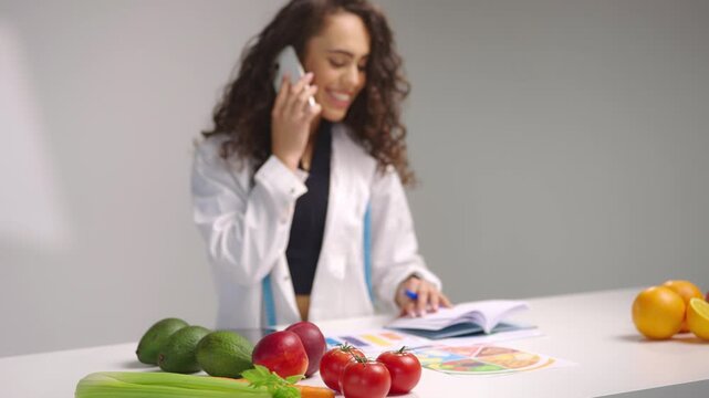 Young female nutritionist smiling while talking on phone and reviewing a calendar at a desk with fresh fruits, vegetables and diet charts, creating healthy meal plans in a professional wellness space.