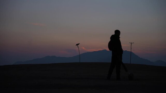 Lone silhouetted cricketer holding bat standing on grassy hill summit against glowing pink and purple twilight sky in remote mountain village