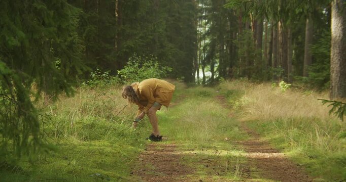 A woman in a yellow jacket bends down to forage in the grass along a forest trail during golden hour. Perfect for sustainability and wellness content.