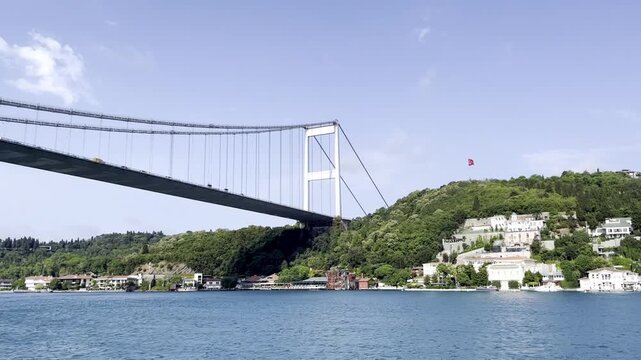 Scenic view of the Fatih Sultan Mehmet Bridge over the Bosporus strait with green hills and traditional houses on the coastline in Istanbul, Turkey